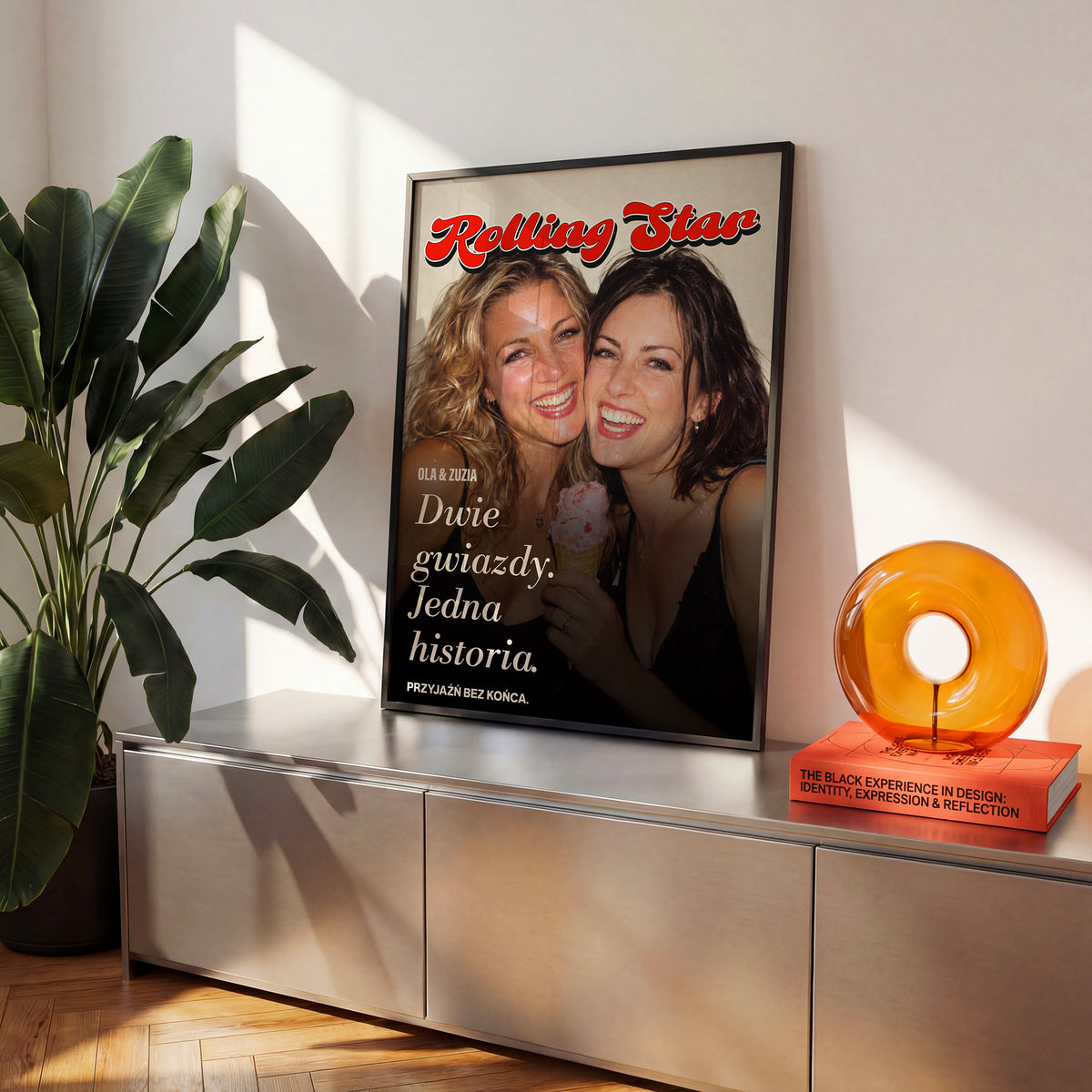 Personalizowany plakat Rolling Star Dwie Gwiazdy w czarnej ramce na komodzie. Czerwony tytuł, wspólne zdjęcie i kursywa tworzą osobistą okładkę ze zdjęciem.