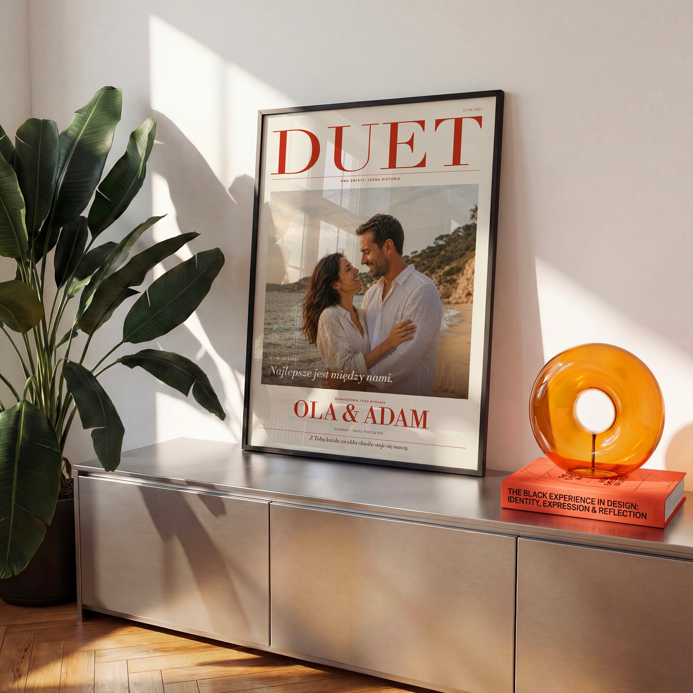 Plakat magazynowy DUET ze zdjęciem pary i imionami w czarnej ramce na komodzie, obok rośliny i pomarańczowej dekoracji | C-PL-COVMAG-5-2130-NF