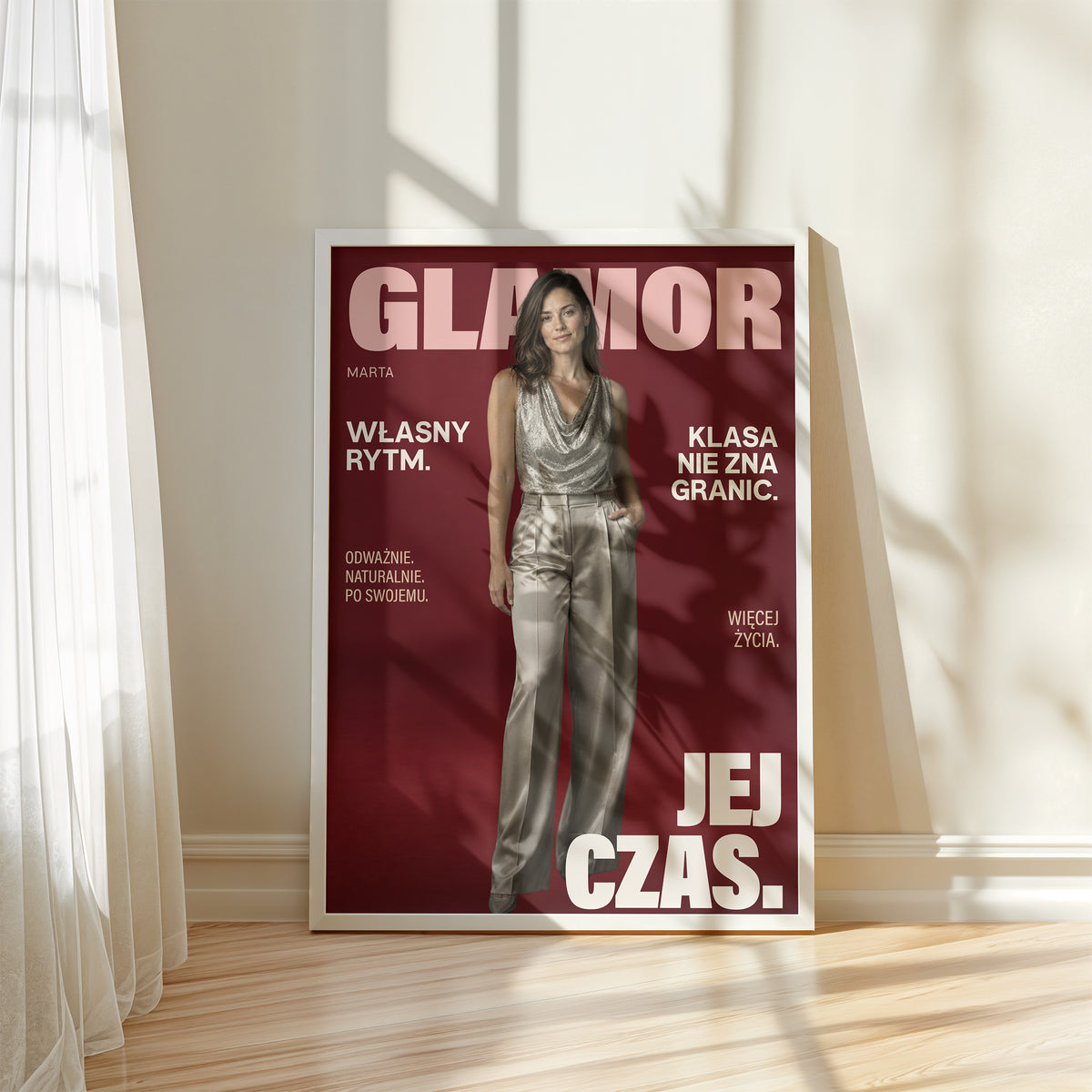 Personalizowany plakat GLAMOR w białej ramce przy oknie. Bordowe tło, portret i jasne napisy na modowej okładce portretowej.