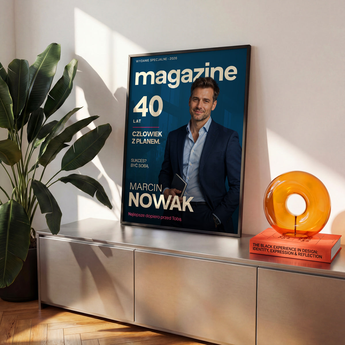 Personalizowany plakat MAGAZINE — CZŁOWIEK Z PLANEM w czarnej ramce ustawiony na komodzie, z własnym portretem, imieniem i napisami na okładce magazynu.