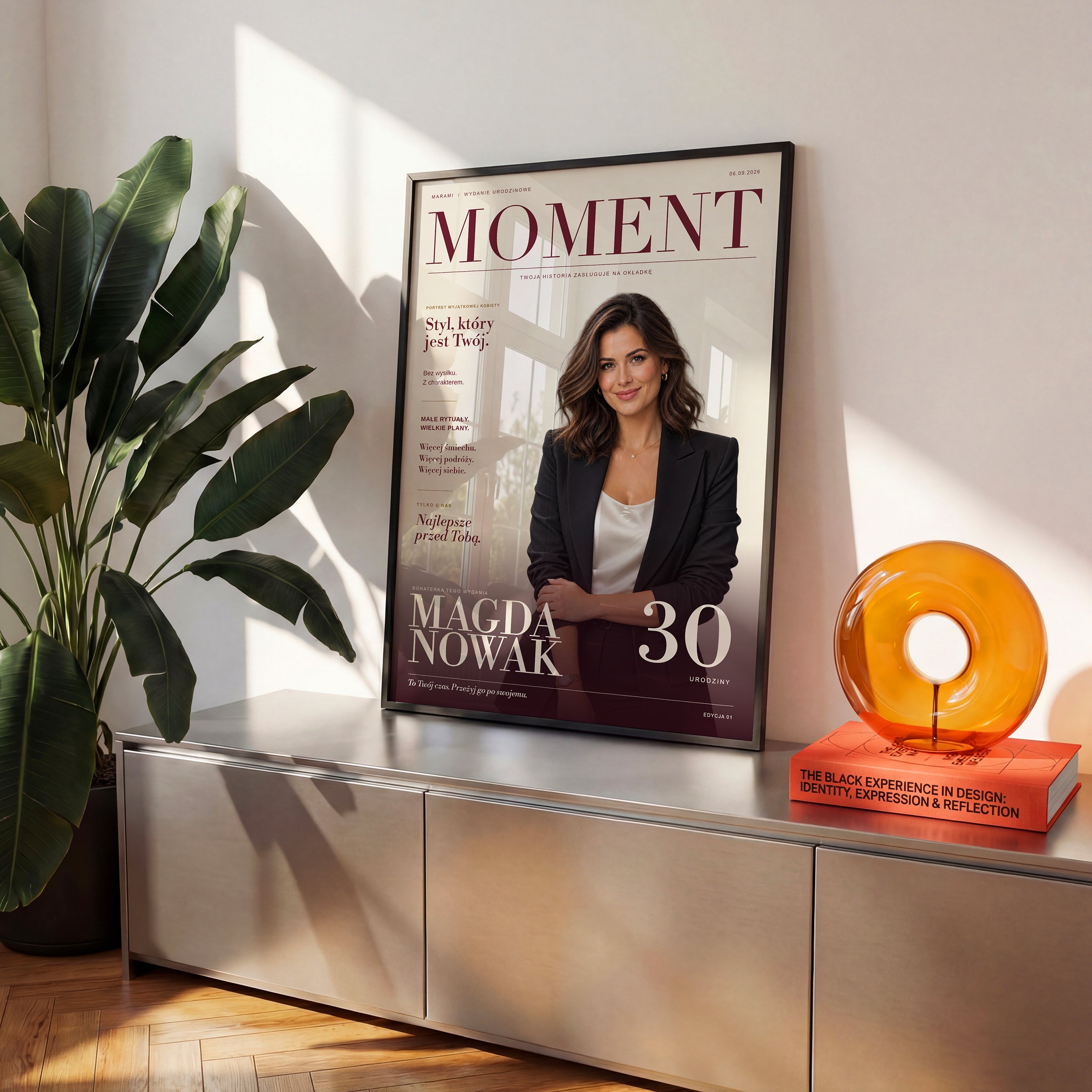 Plakat magazynowy MOMENT w czarnej ramce na komodzie w słonecznym wnętrzu. C-PL-COVMAG-3-2130-NF
