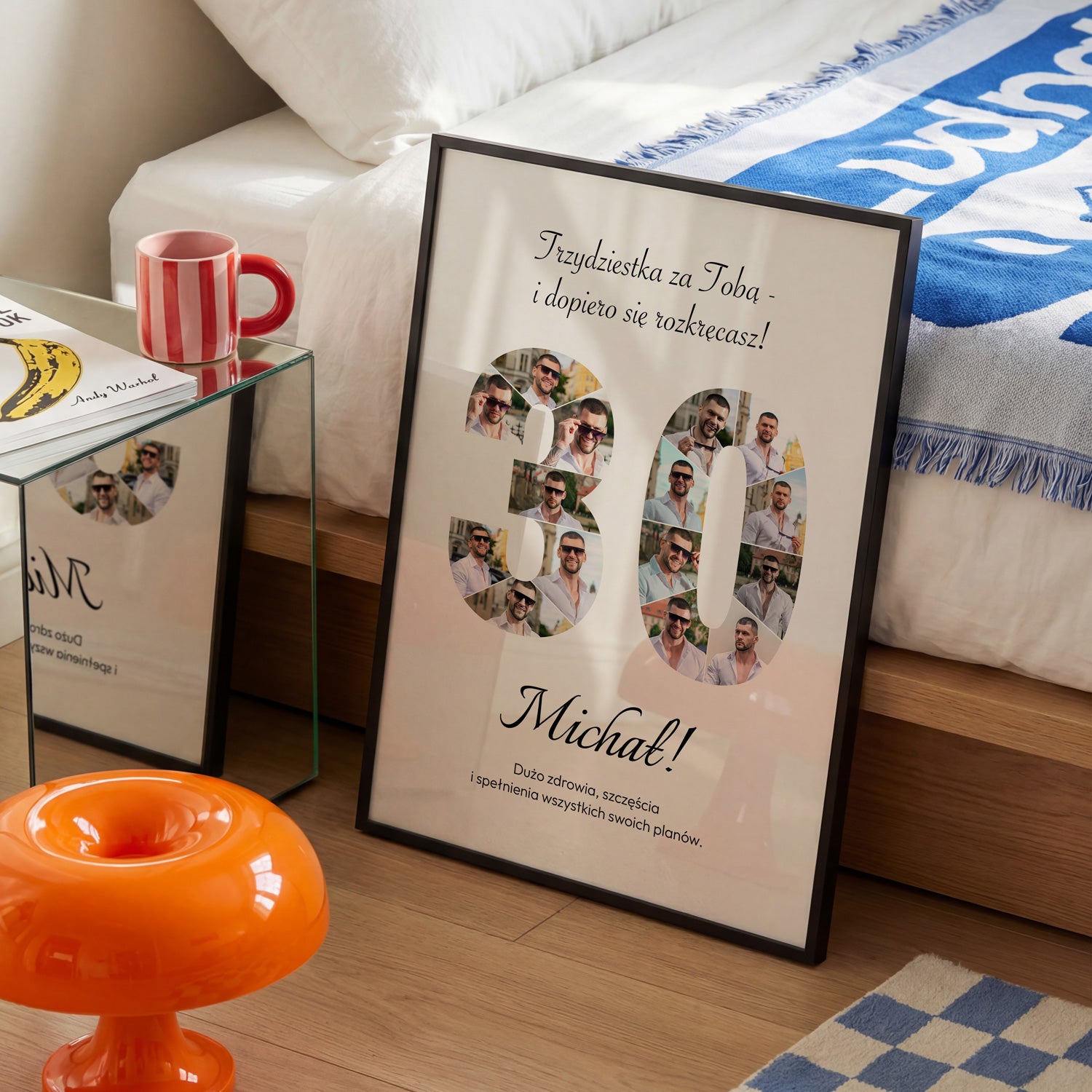 Plakat na 30 urodziny w czarnej ramce z kolażem 16 zdjęć w kształcie cyfry 30 i personalizowaną dedykacją obok łóżka z niebieską narzutą