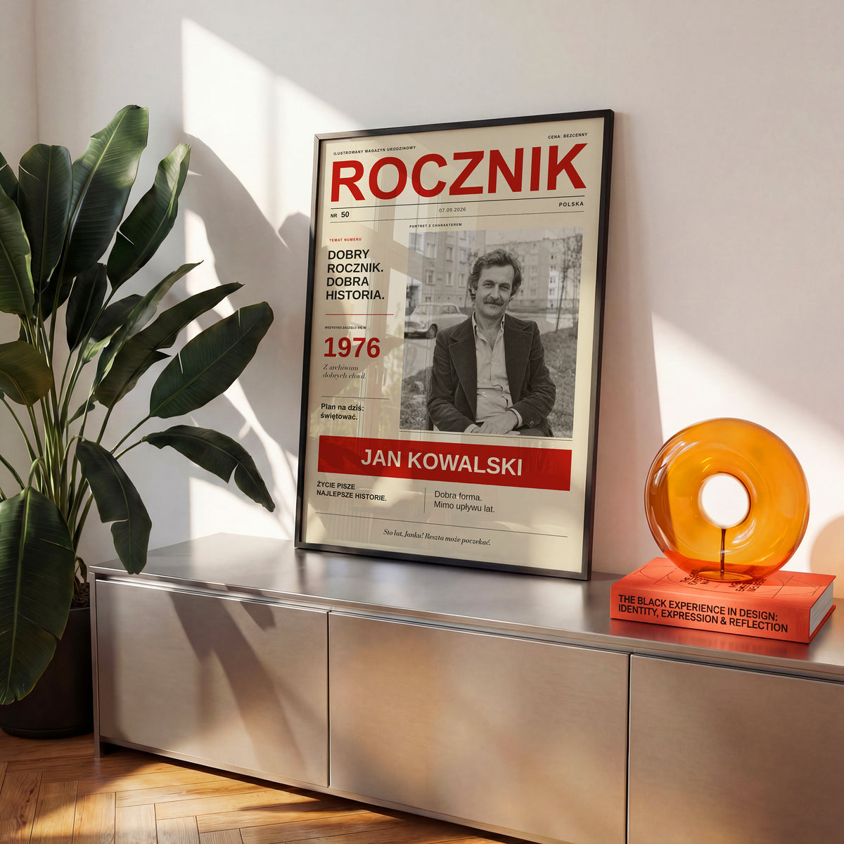Plakat urodzinowy ROCZNIK w czarnej ramce na komodzie obok rośliny, okładka retro z czarno-białym zdjęciem i czerwonym tytułem | C-PL-COVMAG-6-2130-NF