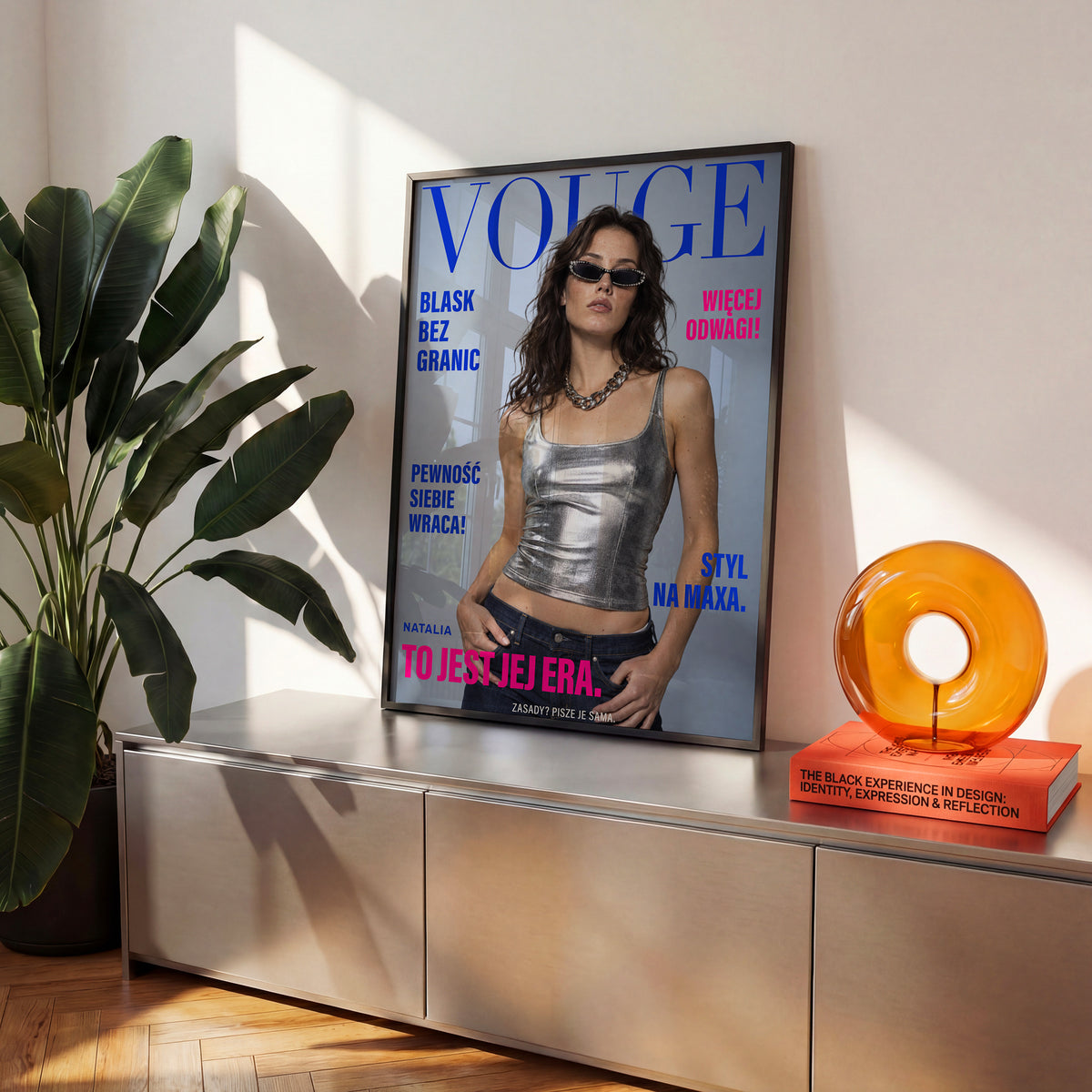 Personalizowany plakat Vouge Jej Era w czarnej ramce na komodzie. Kobaltowy tytuł, różowe hasła i portret na modowej okładce portretowej.