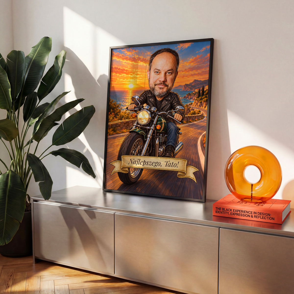 Motocyklista — plakat w czarnej ramie na komodzie we wnętrzu, z karykaturą mężczyzny na motocyklu, na nadmorskiej drodze o zachodzie słońca. C-PL-KAR-06-2130-NF